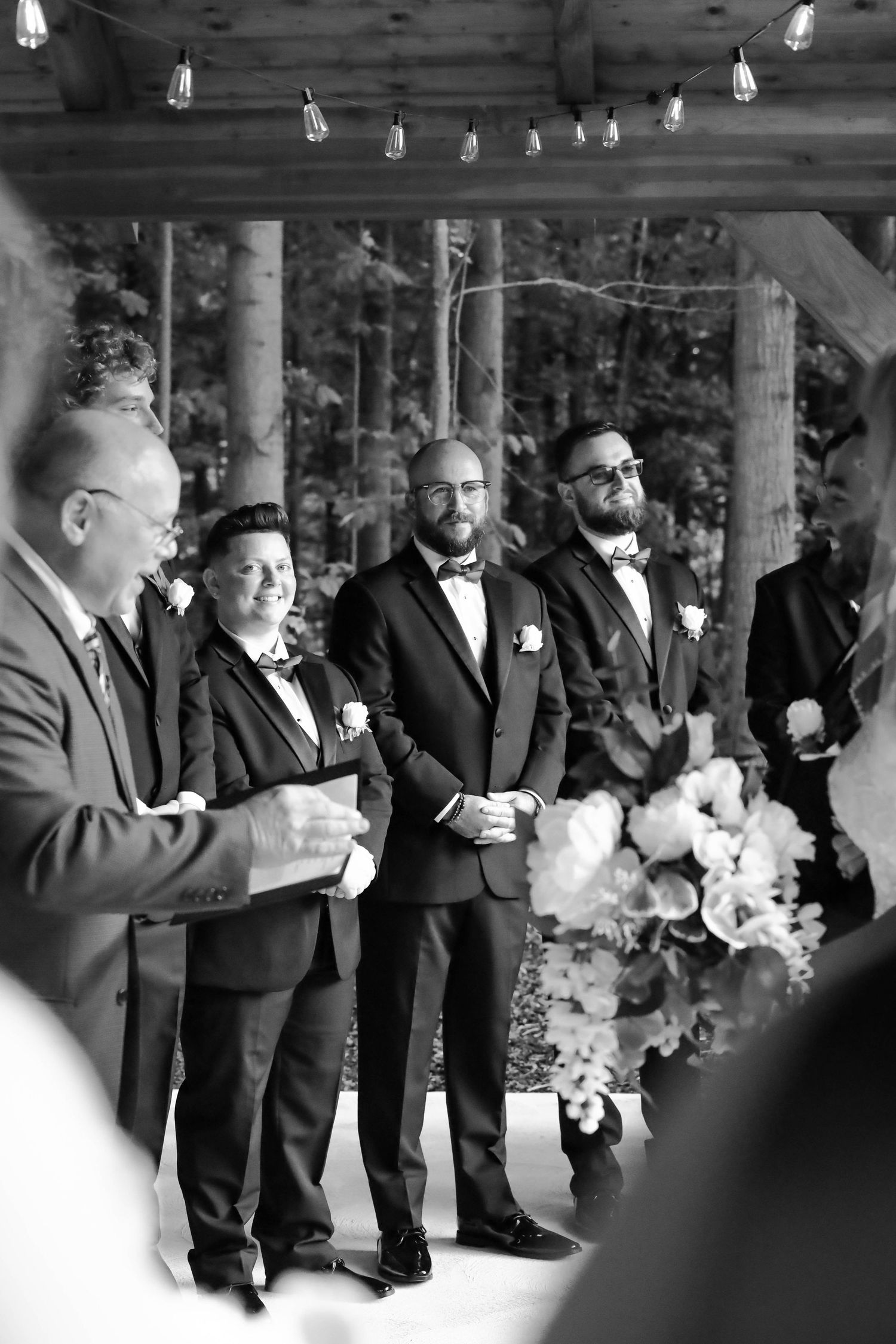 Black and white photo of groomsmen standing in rustic covered venue with string lights overhead.