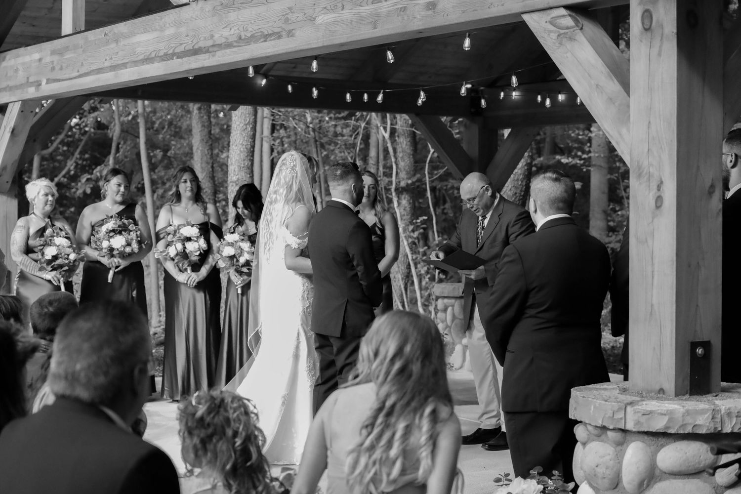 Black and white photo of outdoor wedding ceremony under wooden pavilion with string lights.
