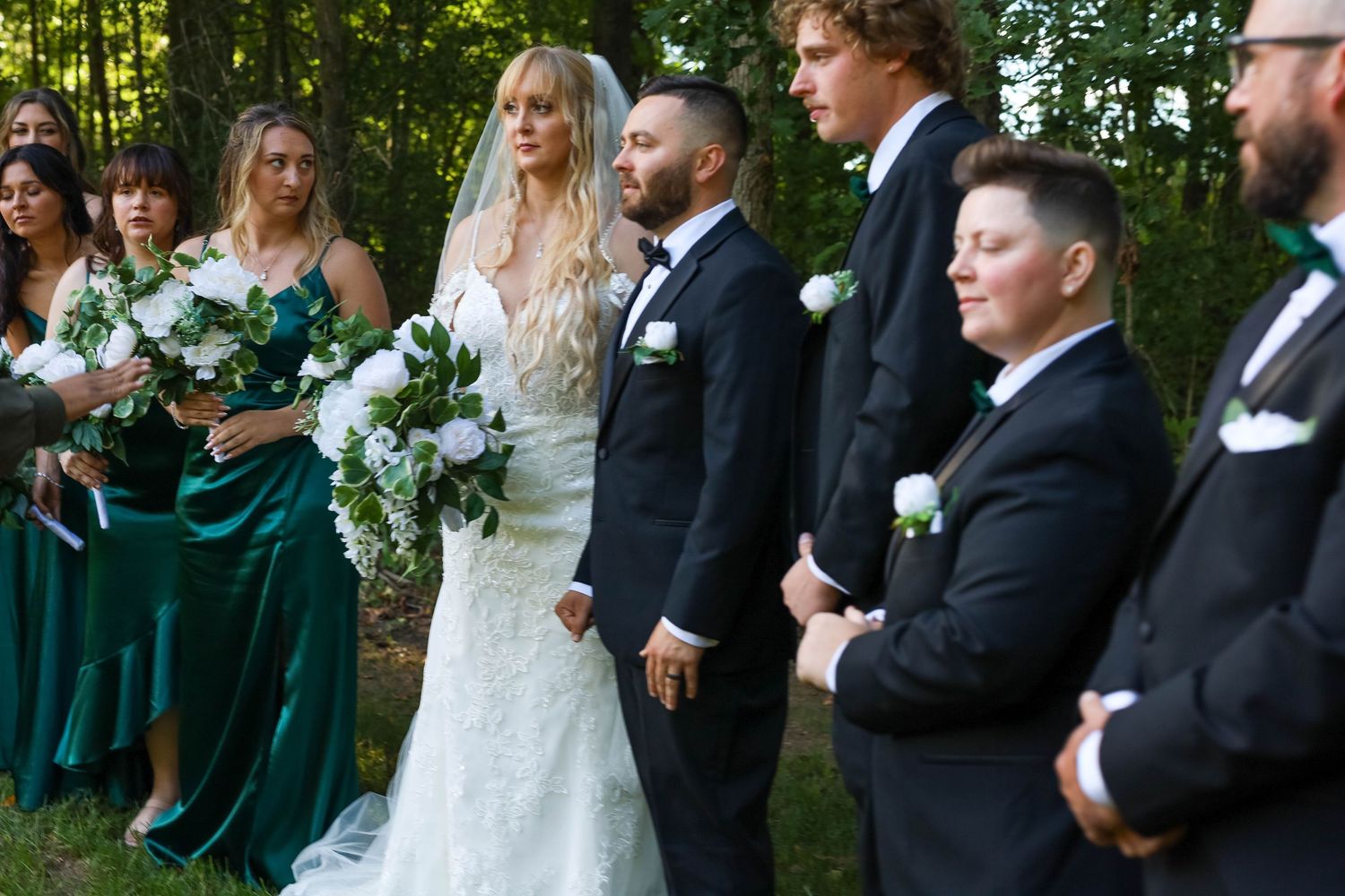Wedding ceremony guests in formal black attire and green dresses gather outdoors amidst trees and sunlight.