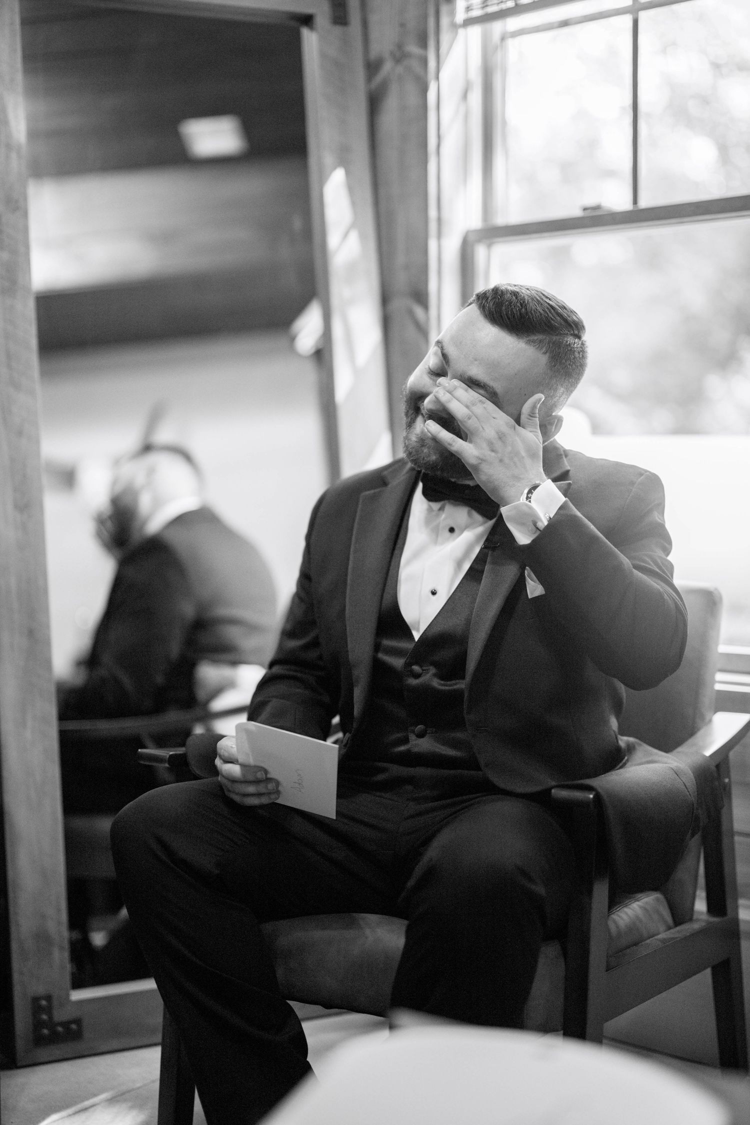 Emotional groom in black tuxedo sits alone covering face with hands in black and white photography.