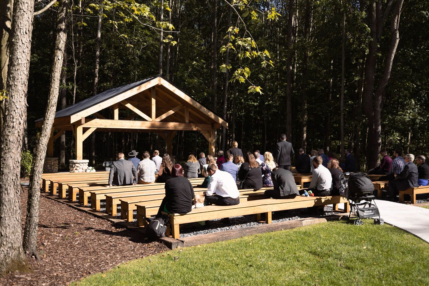 Rustic outdoor wedding venue with wooden pavilion and bench seating surrounded by forest.