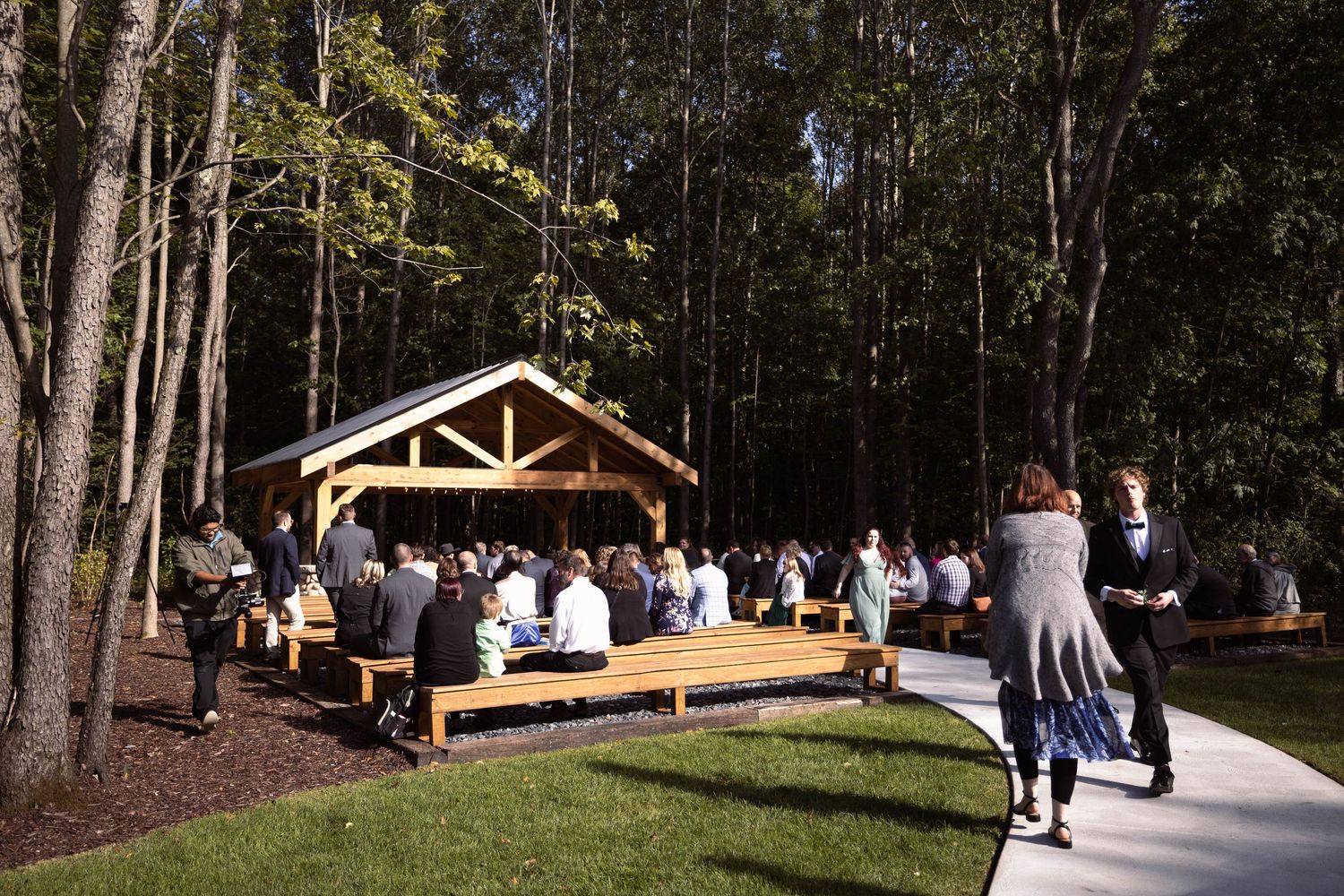 Outdoor wedding ceremony venue with wooden pavilion surrounded by forest as guests gather on benches.