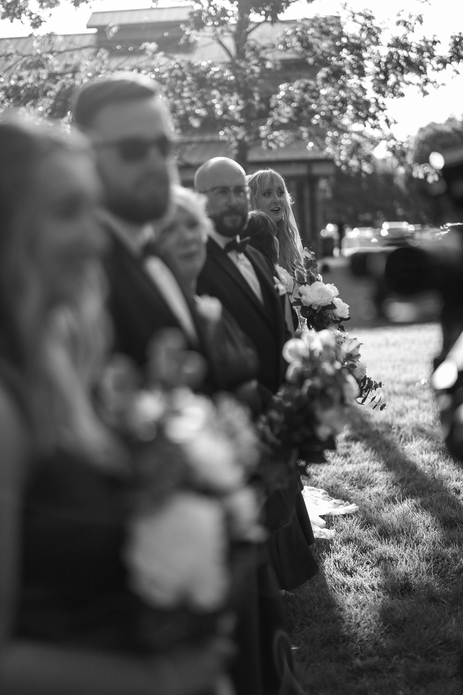 Black and white wedding photo showing groomsmen in formal attire standing in a line with flower bouquets at an outdoor ceremony.