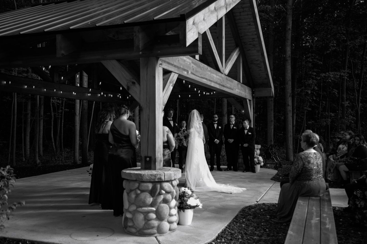 Black and white photo of a wedding ceremony taking place at a rustic outdoor venue with stone pillars and wooden beams.