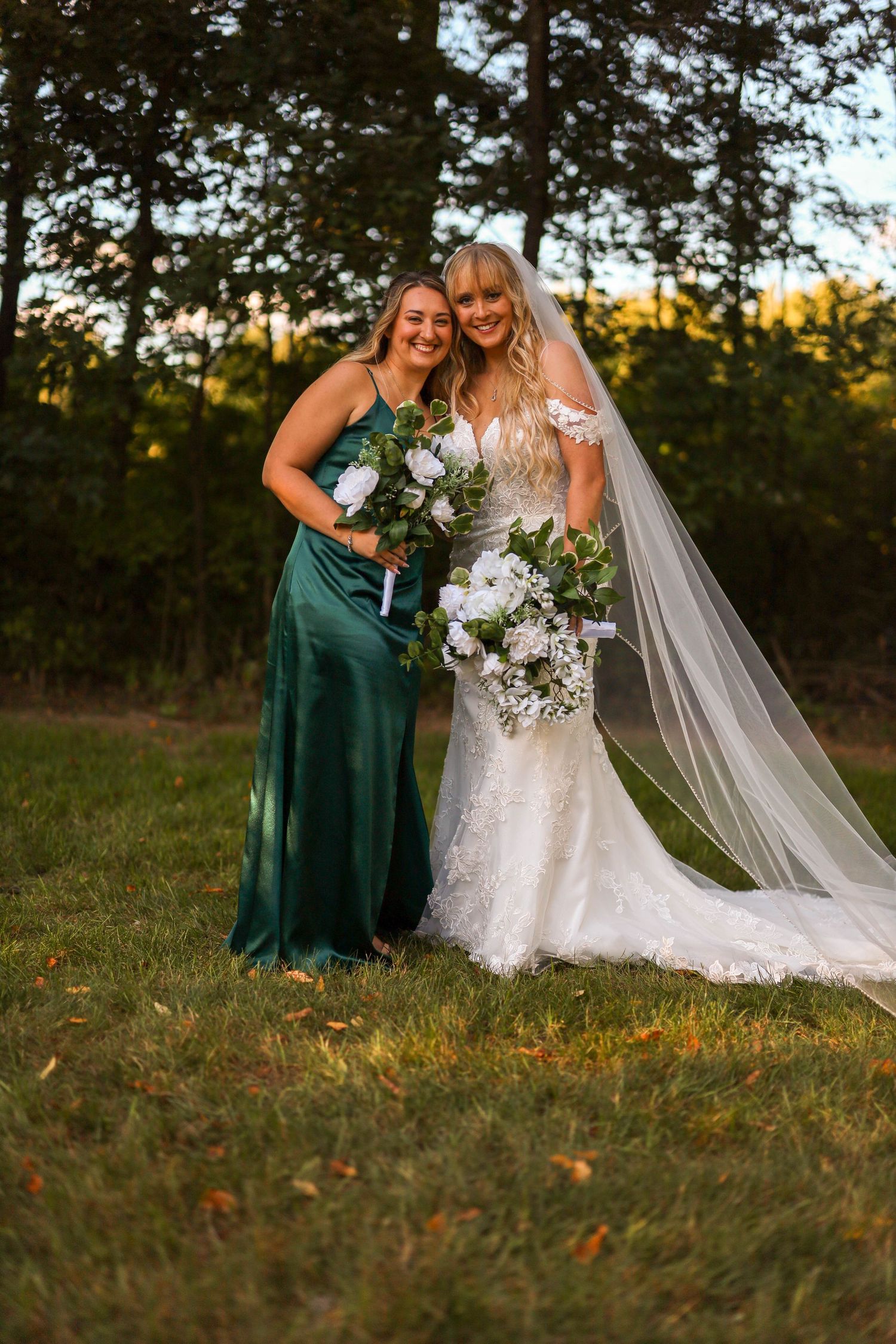 Bride in lace gown and bridesmaid in emerald dress pose with white bouquets during sunset outdoor wedding photography.