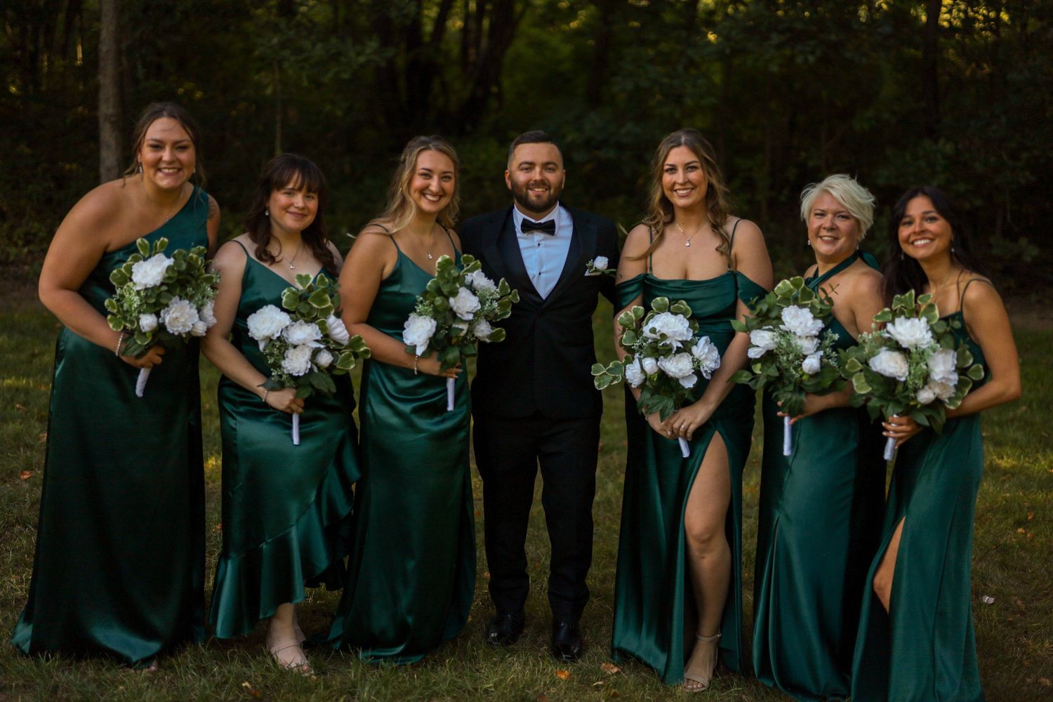 Wedding party in emerald green dresses poses together holding white floral bouquets at an outdoor venue.