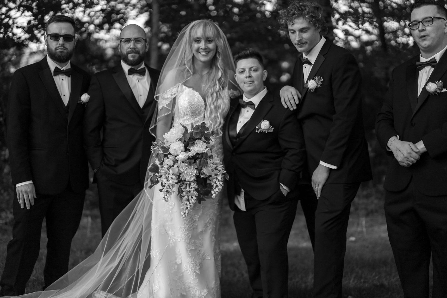 Members of wedding party stand together outdoors in formal black and white attire for group photos.