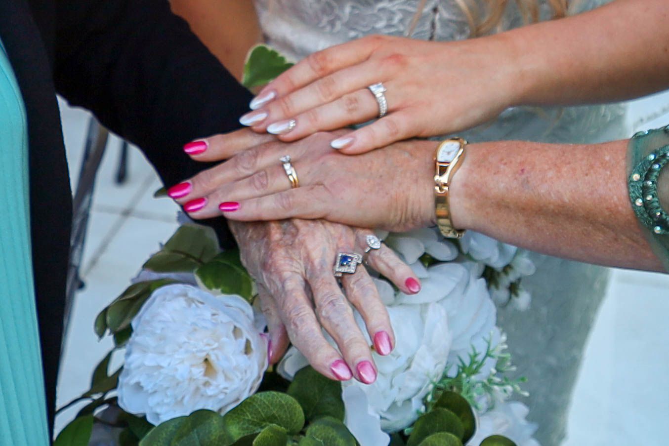Three hands with rings and pink manicures rest together on white flowers.