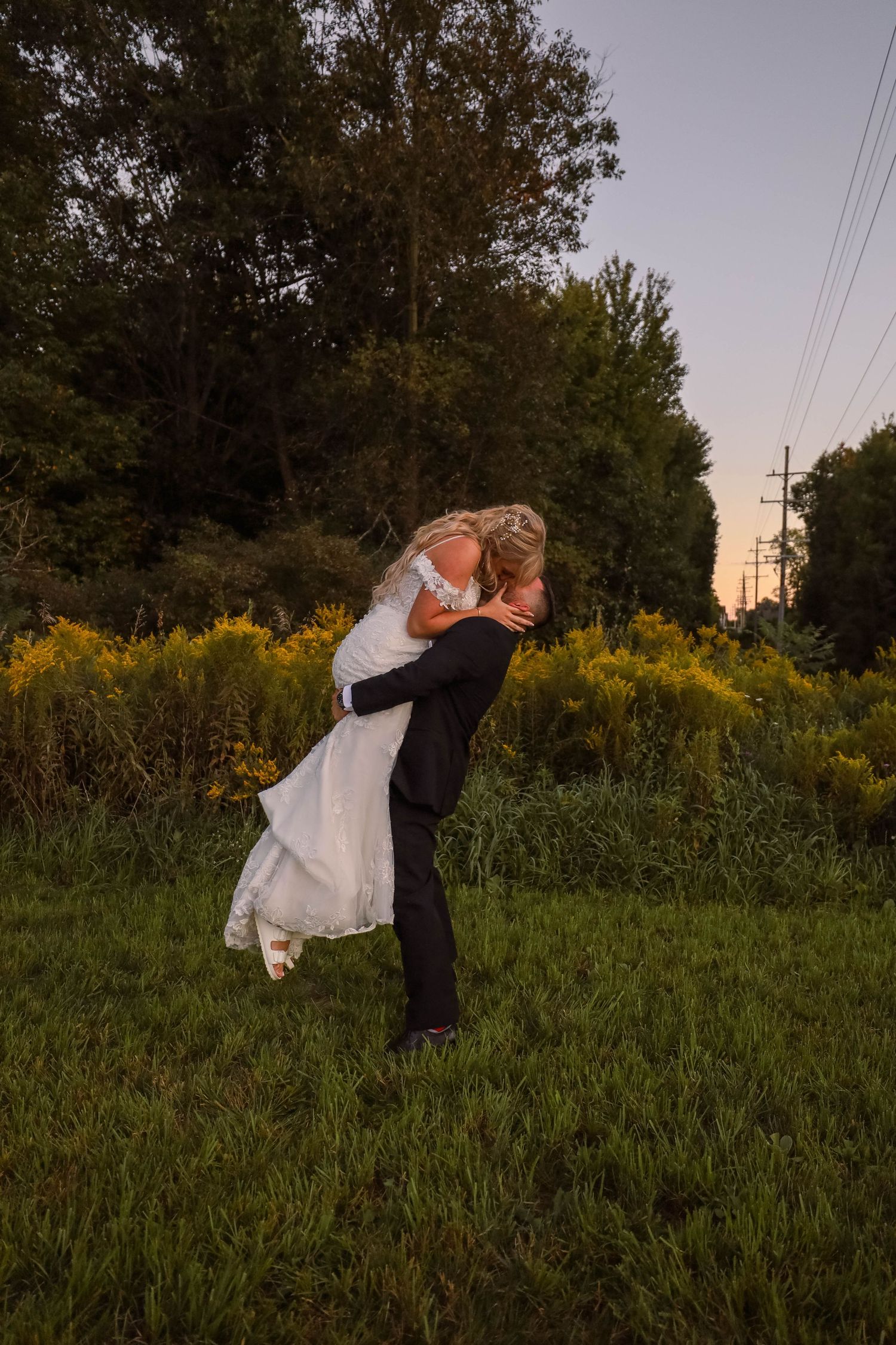 A romantic sunset wedding photo of a couple embracing in a meadow with golden wildflowers and trees.