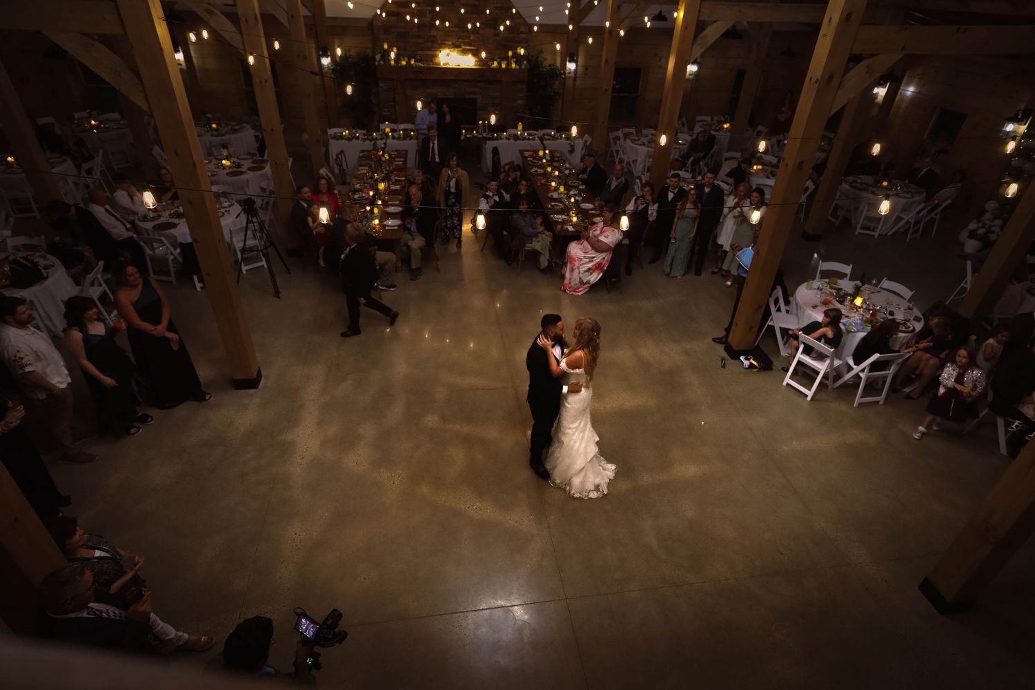 A couple shares their first dance at a rustic indoor wedding reception while guests watch from their seats around the room.