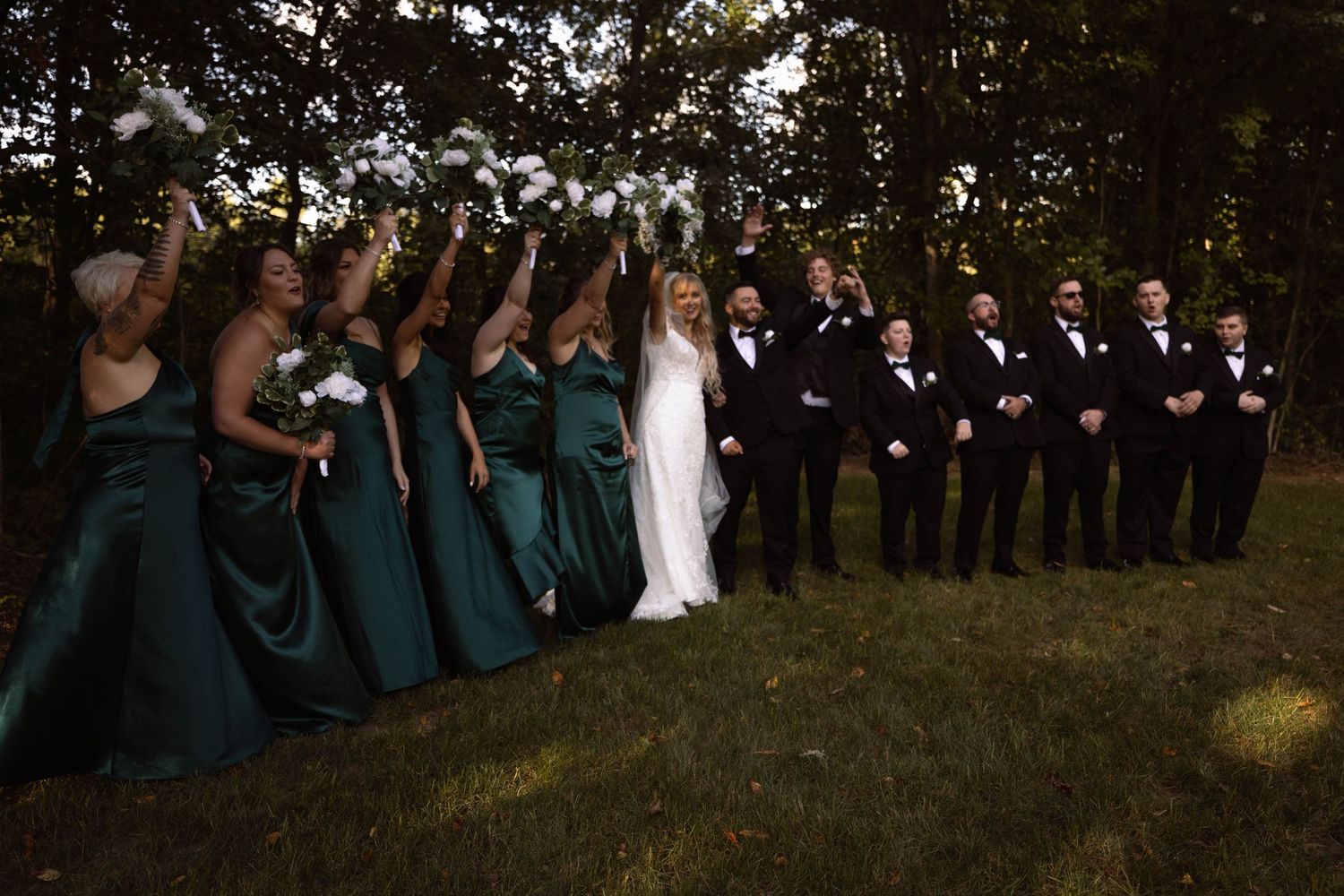 Wedding party stands in a row outdoors with bridesmaids in dark green dresses and groomsmen in black suits.