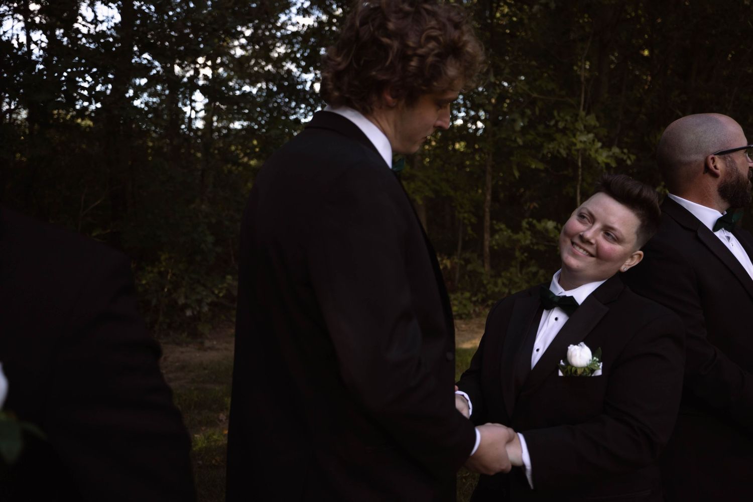 Two people shake hands during an outdoor wedding ceremony in a wooded setting at dusk in formal attire.