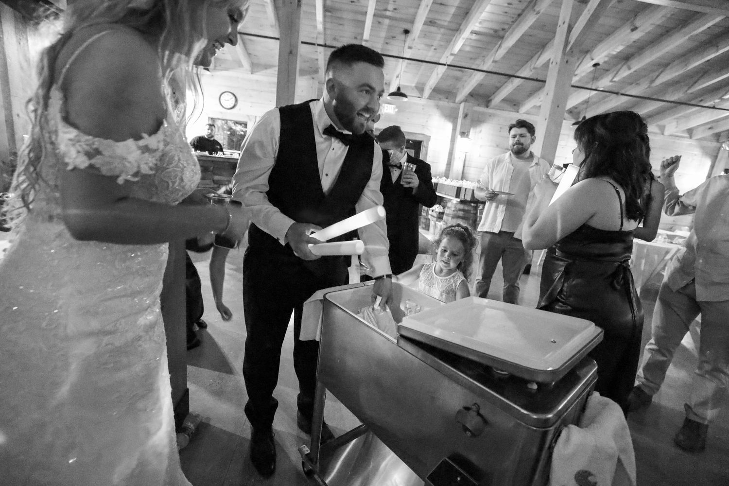 A person in a vest serves refreshments from a vintage suitcase bar setup at an indoor wedding reception.
