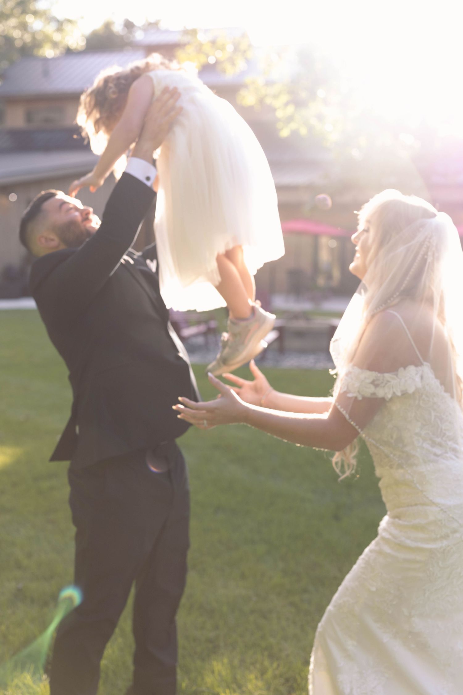 A playful wedding moment in golden sunlight as a couple lifts a small child during outdoor celebrations.