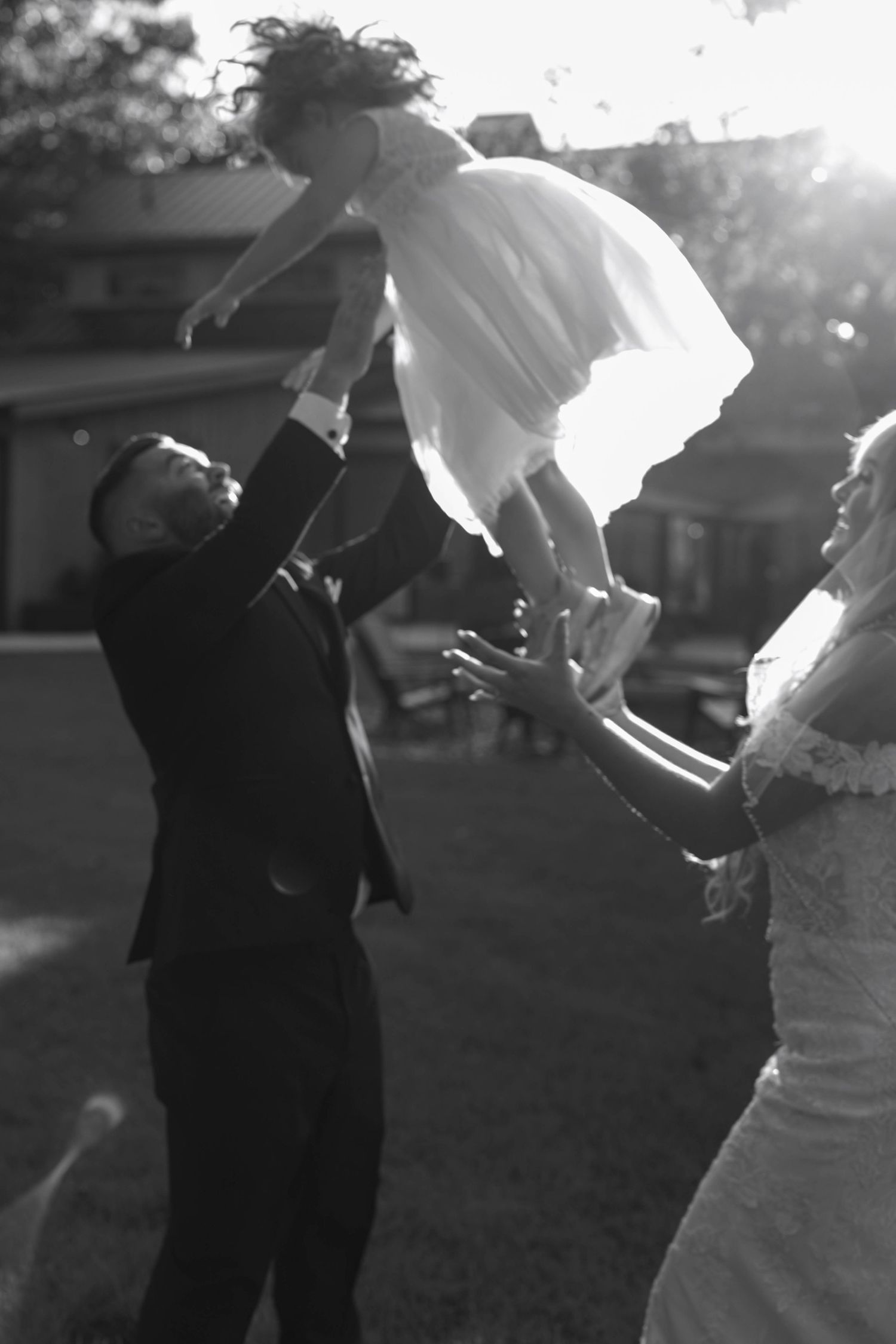 A joyful black and white wedding photo captures a couple playfully tossing a child in a dress during outdoor celebrations.