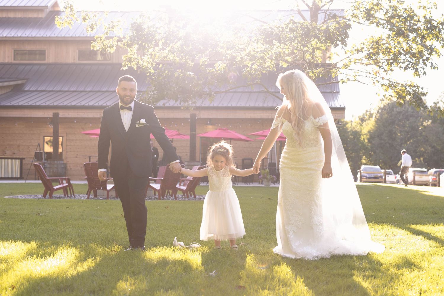 Wedding party walks together on sun-drenched lawn with couple holding hands with flower girl in white dress.