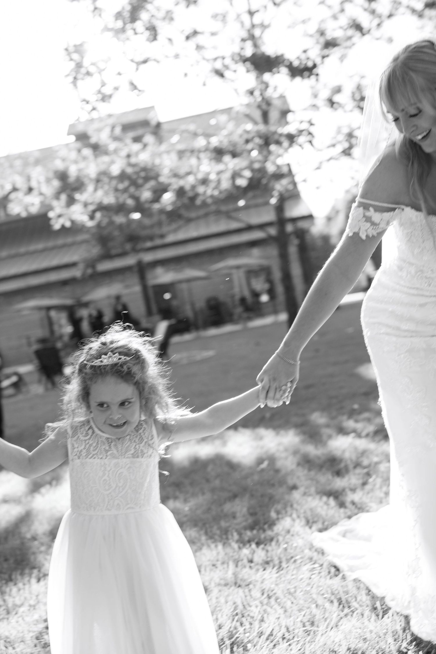 A bride in a white dress holds hands with a young flower girl during an outdoor wedding celebration.