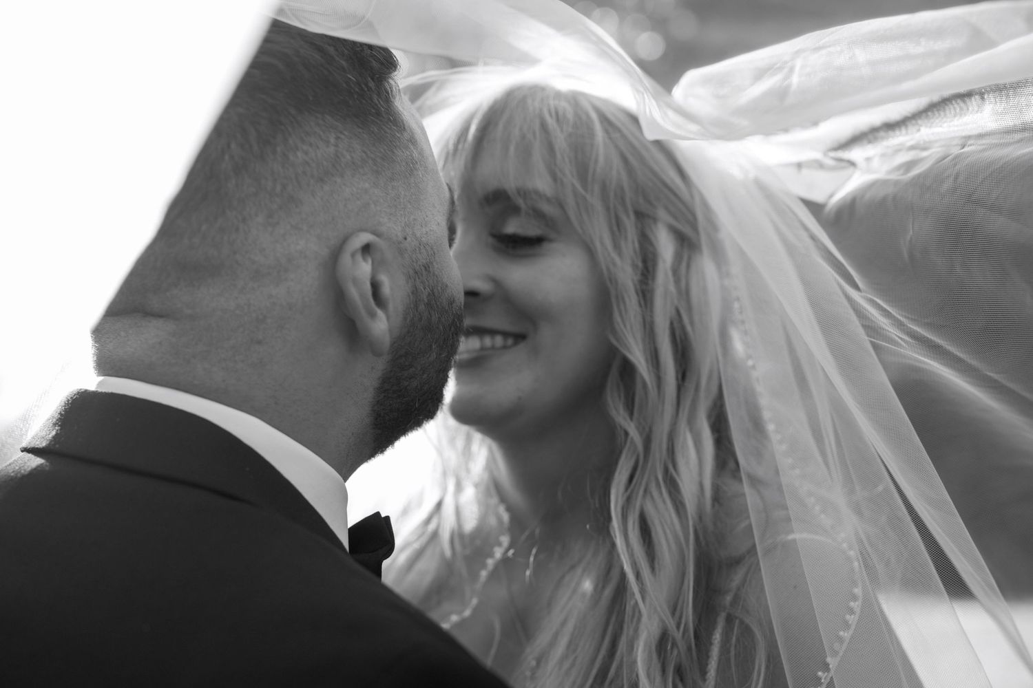 A tender black and white wedding moment captured between a bride and groom as her veil flows in the breeze.