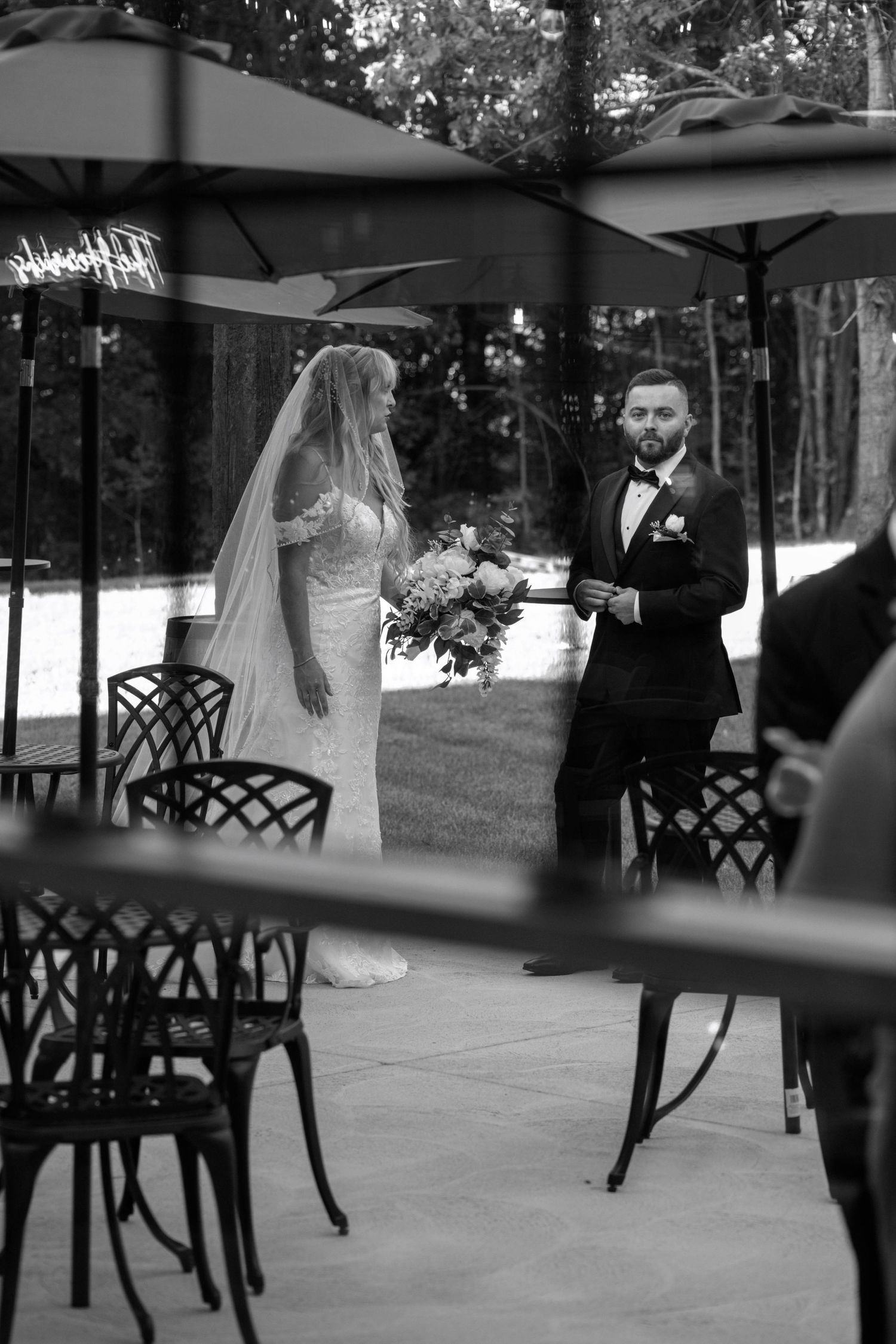 Black and white wedding scene with couple walking between outdoor patio furniture.