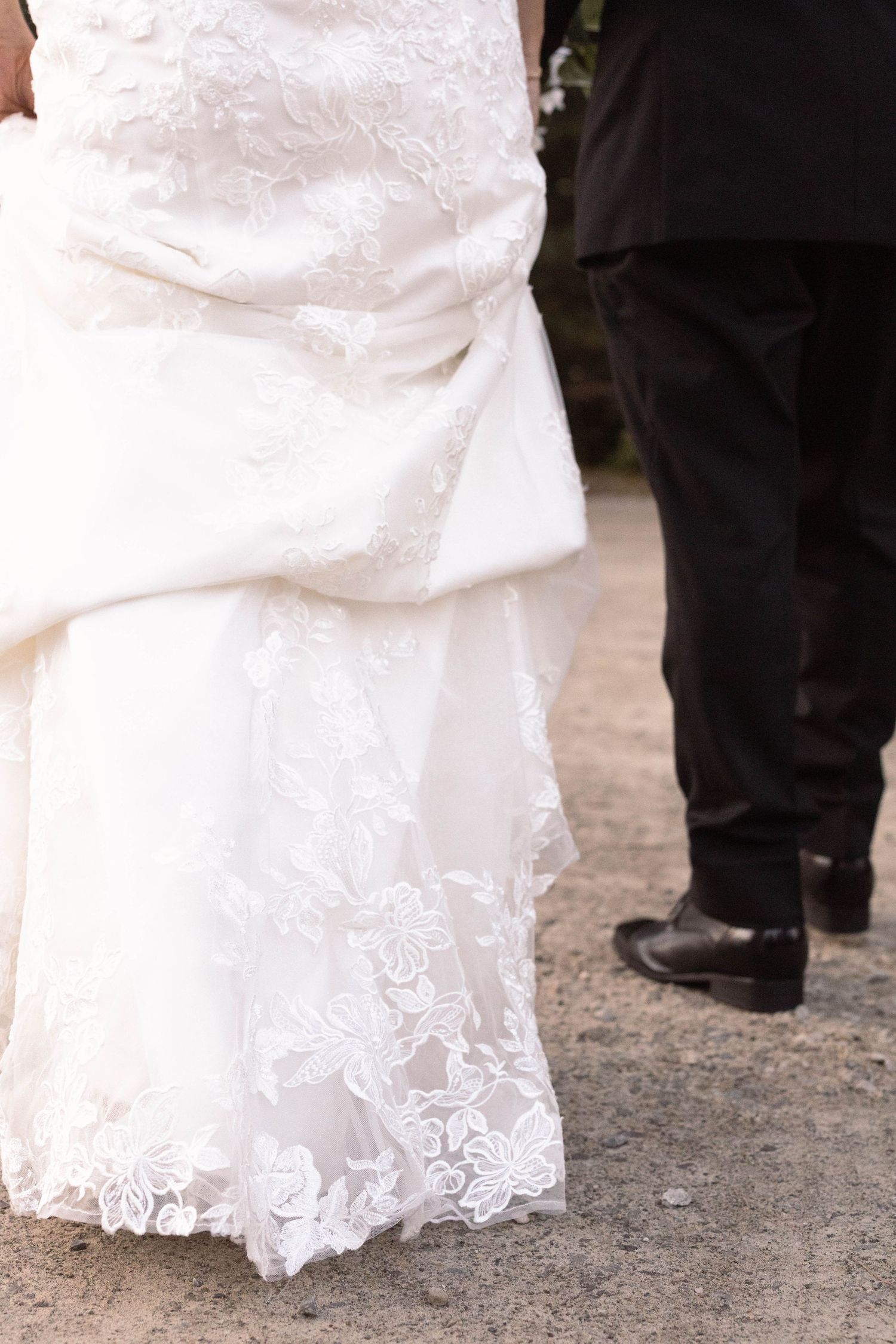 Bottom portion of a white lace wedding dress with intricate embroidery detail visible against dark shoes.