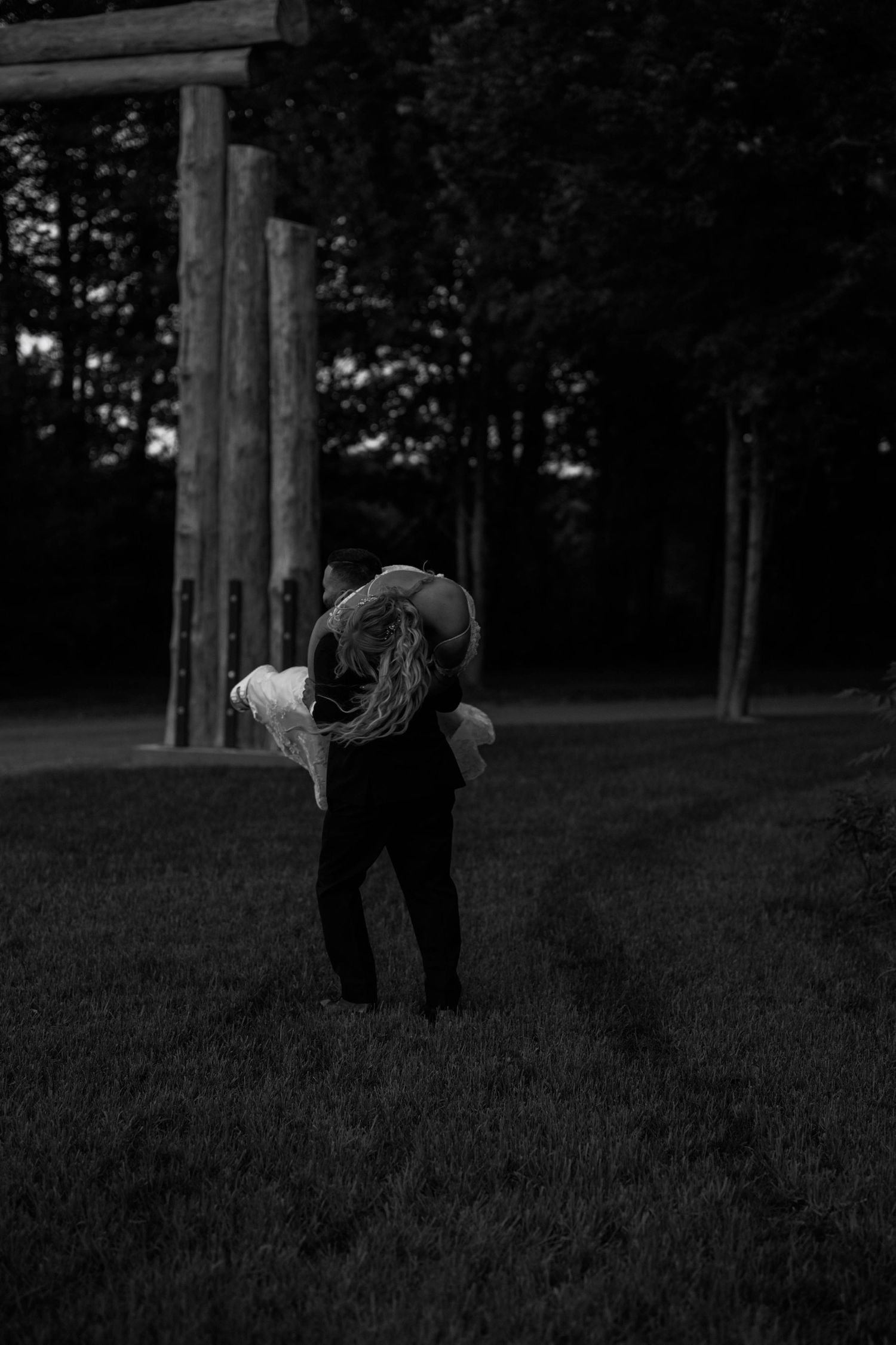 A romantic black and white photo of a couple embracing in front of wooden posts at dusk.