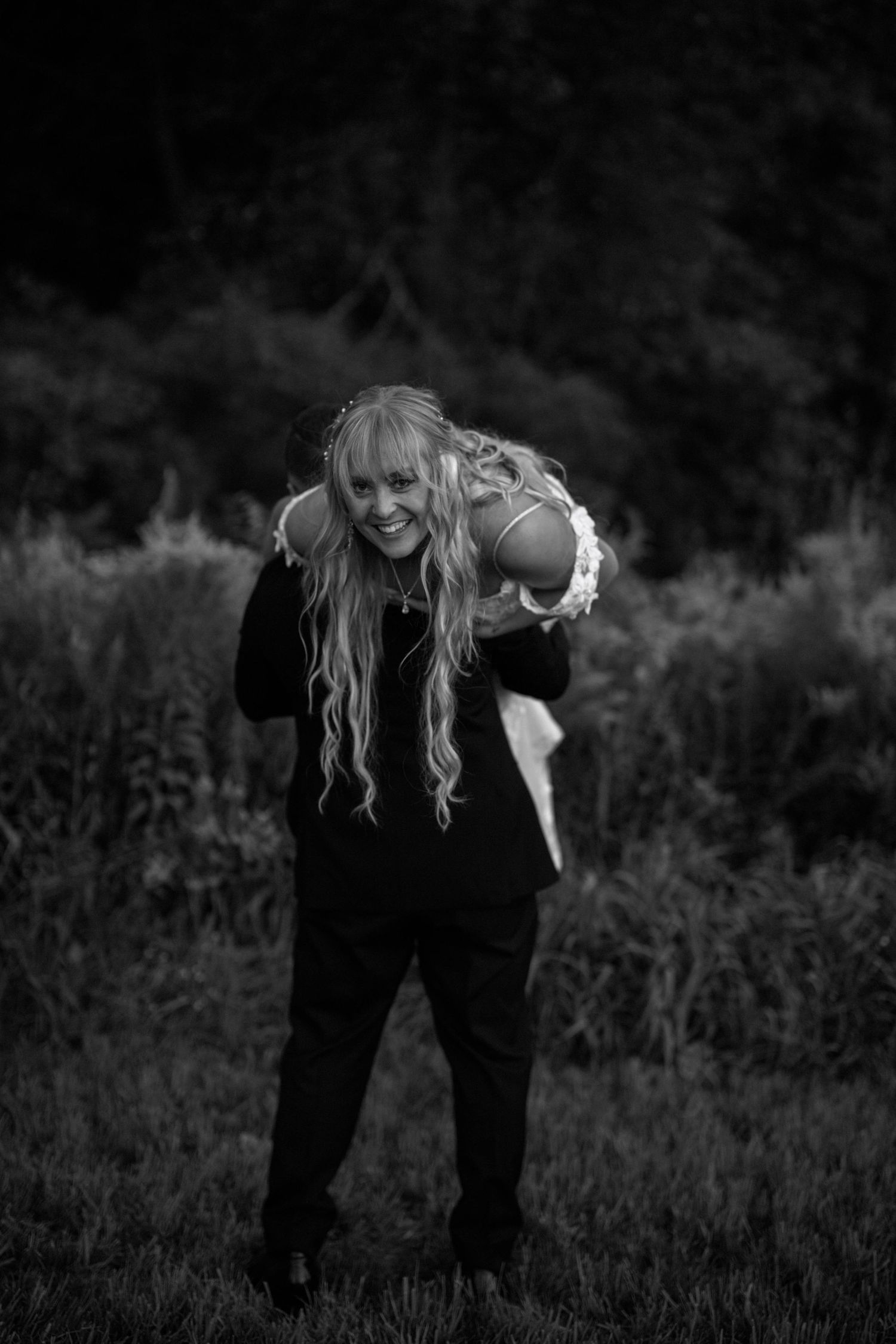 A joyful black and white portrait of a bride laughing while being carried in an outdoor setting.