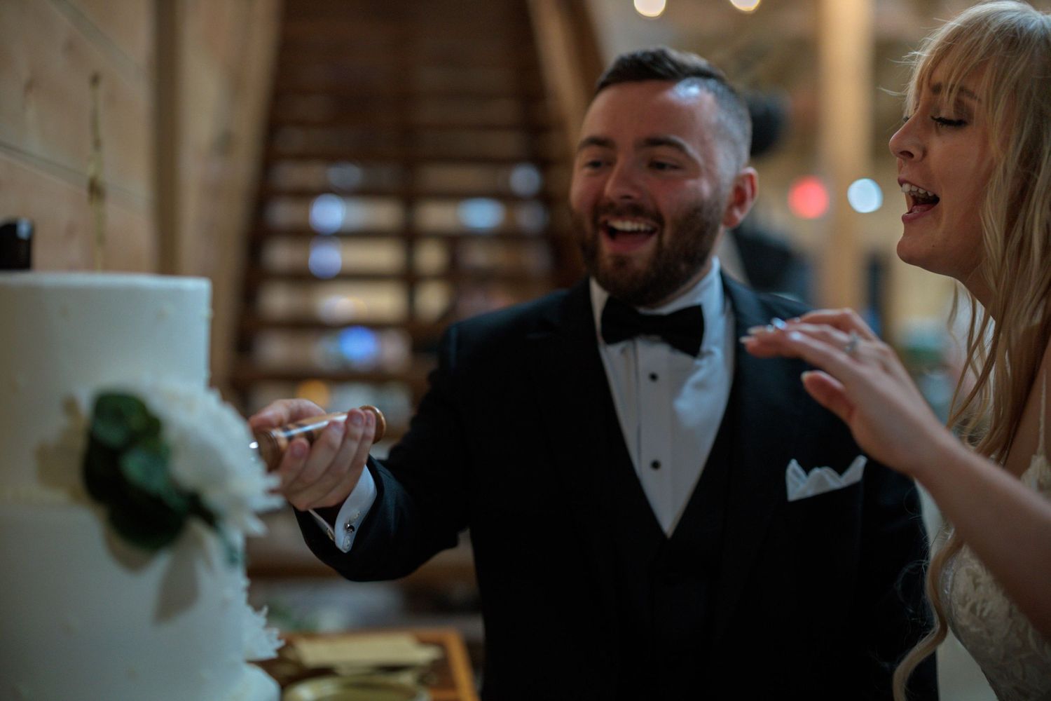 Couple laughing while cutting their wedding cake together at a dimly lit indoor reception.