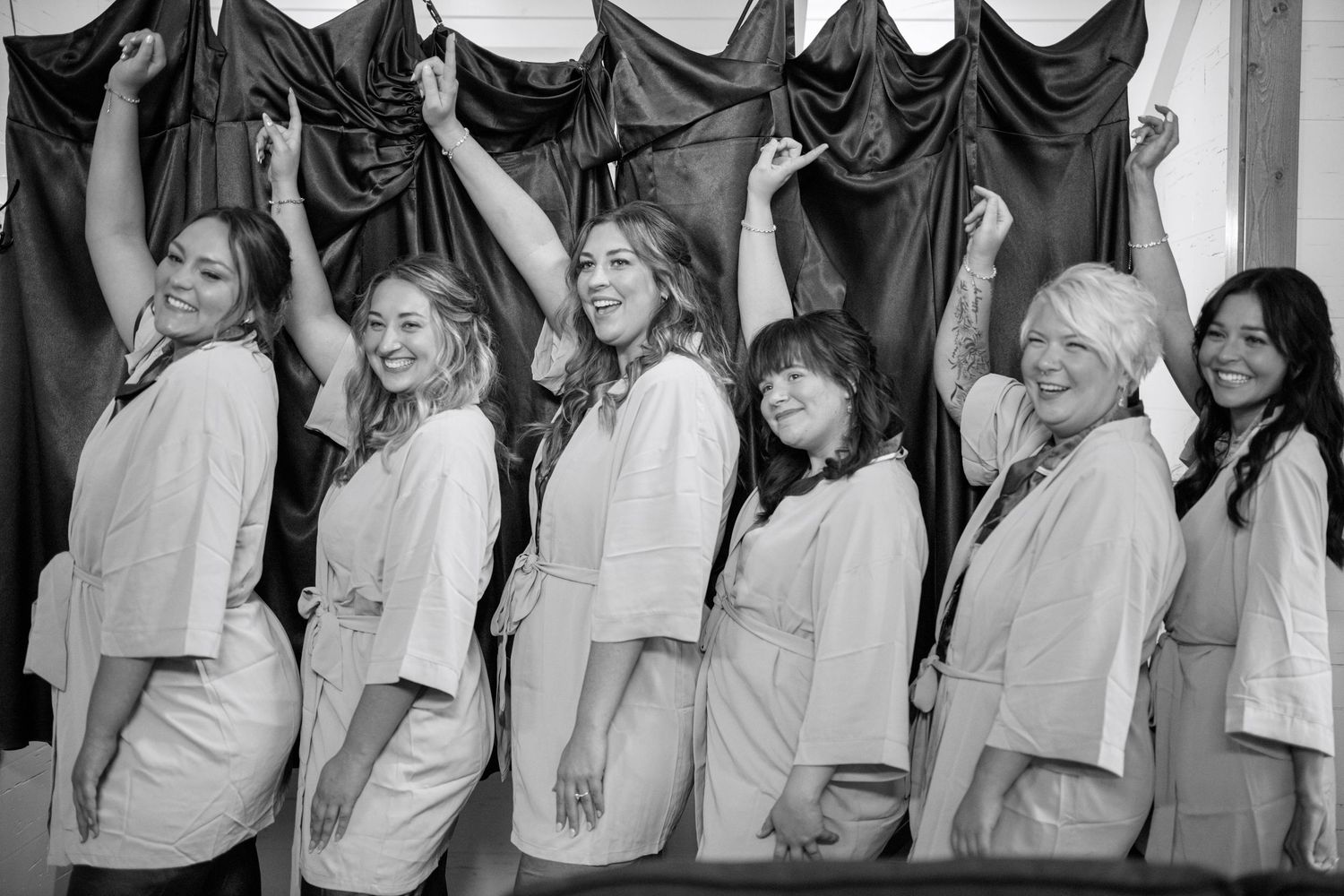 A black and white photograph shows a row of people in light colored outfits posing against draped fabric.