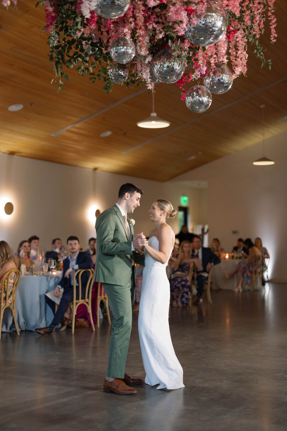 Wedding couple dancing gracefully under pink floral decorations and disco balls during their first dance at a reception venue.