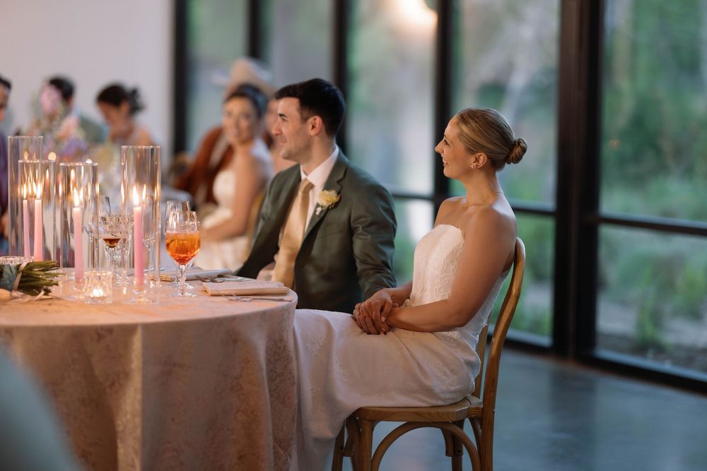 Wedding guests seated at an elegant reception table with glowing candles and soft evening lighting through windows.