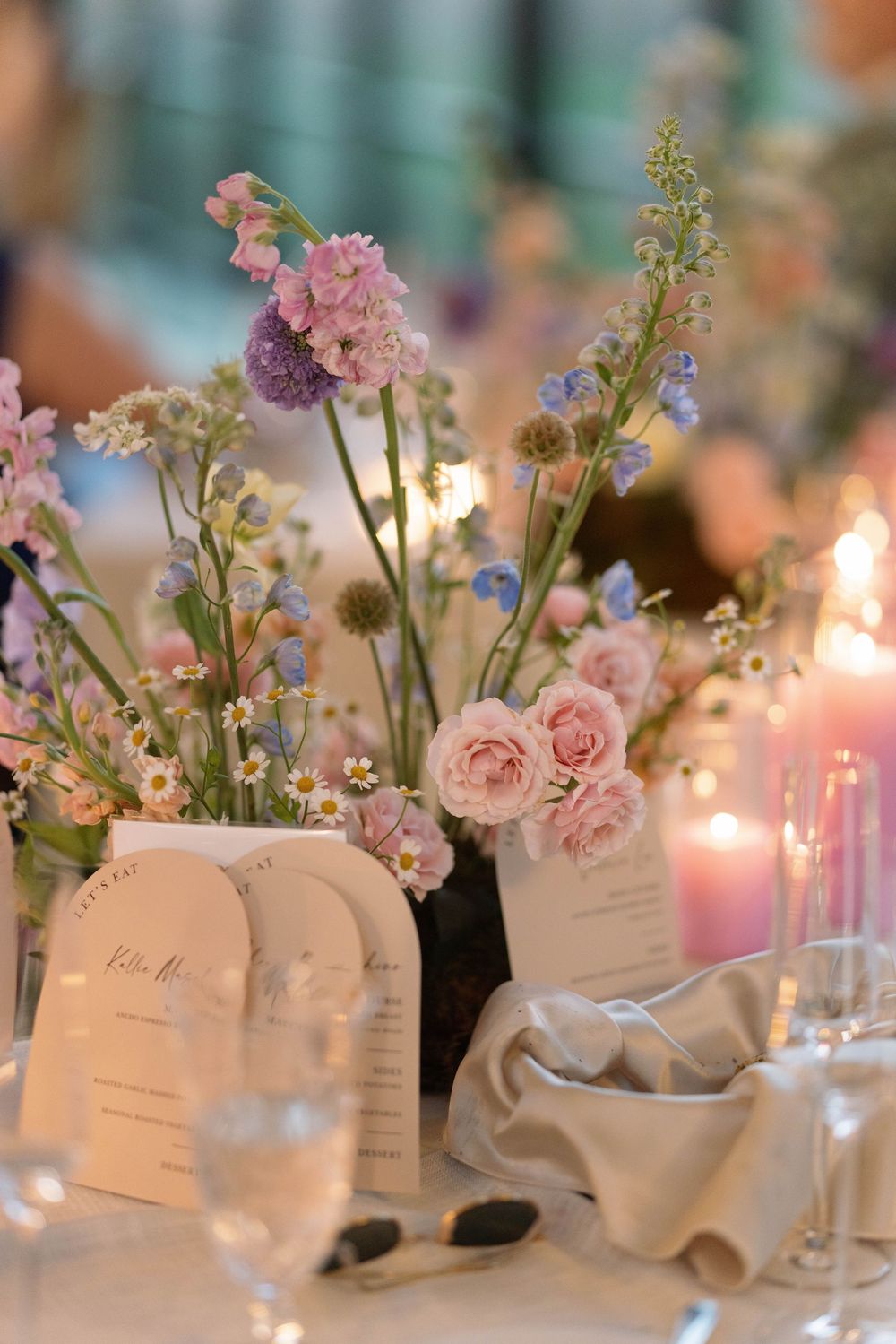 Elegant wedding table centerpiece features pink roses and wildflowers in white ceramic vases with candlelight and place cards.