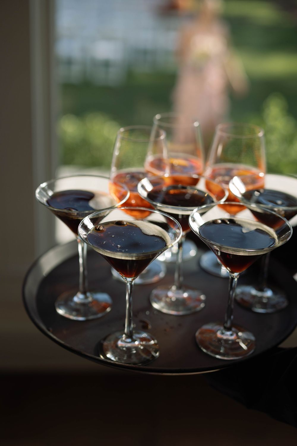Martini cocktails served on dark round tray with orange garnishes illuminated by natural window light from side.