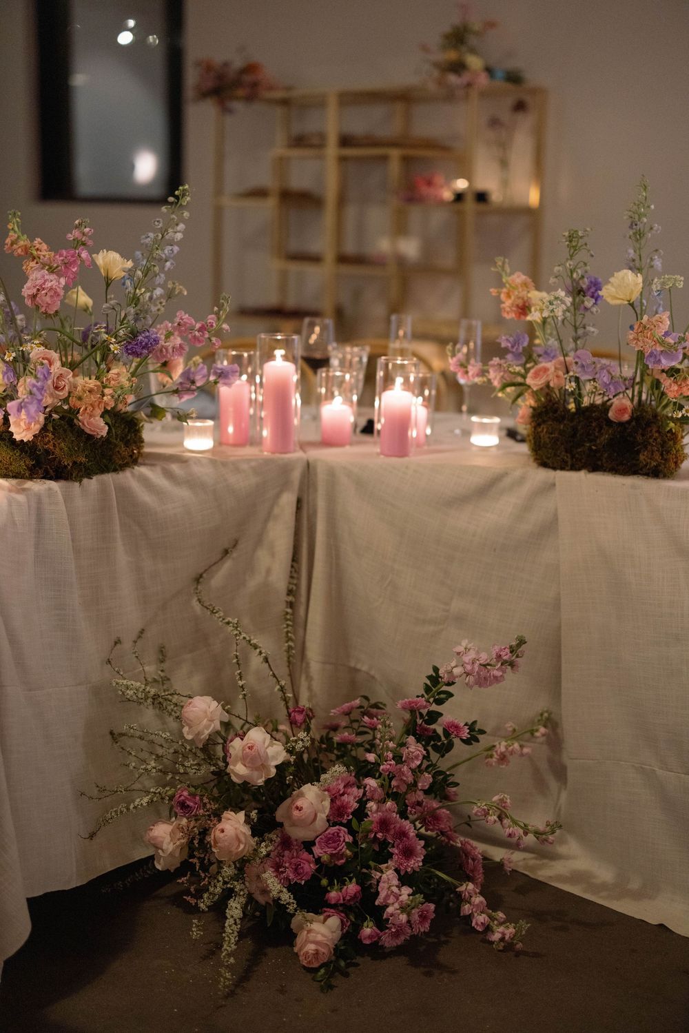 Romantic table setting with pink and purple floral arrangements and glowing candles on white linen tablecloth.