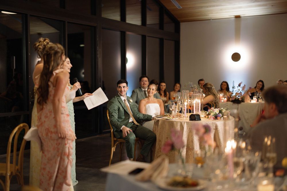 Wedding reception attendees gather in an elegant venue with candlelit tables as a speaker addresses the room.
