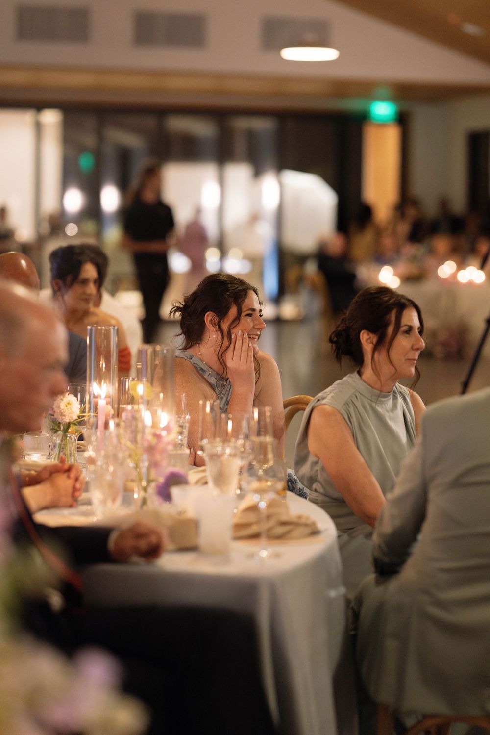 Wedding reception tables with candlelit centerpieces and white tablecloths in a dimly lit venue.