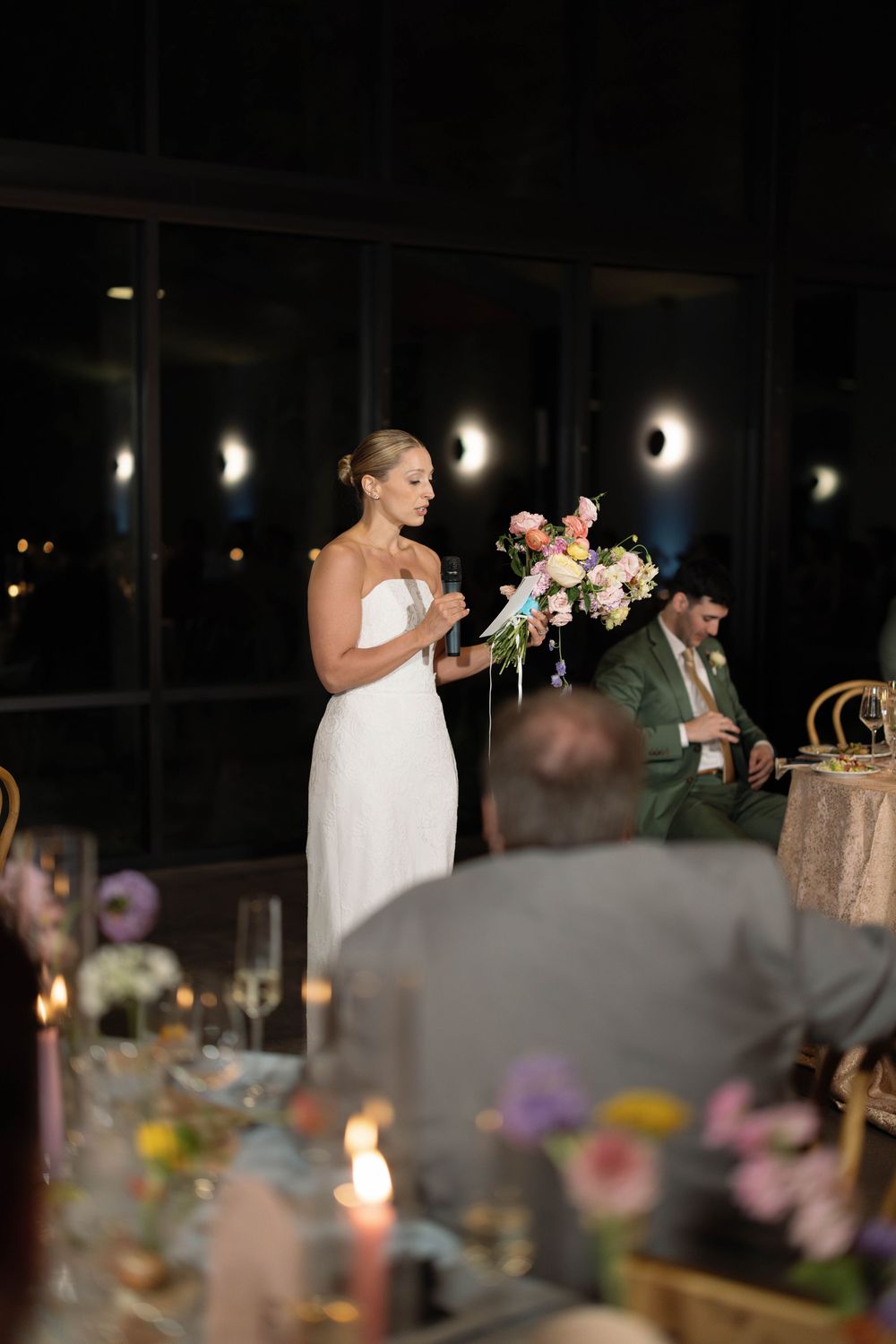 A wedding reception with the bride giving a speech while holding a colorful floral bouquet in a dimly lit venue.