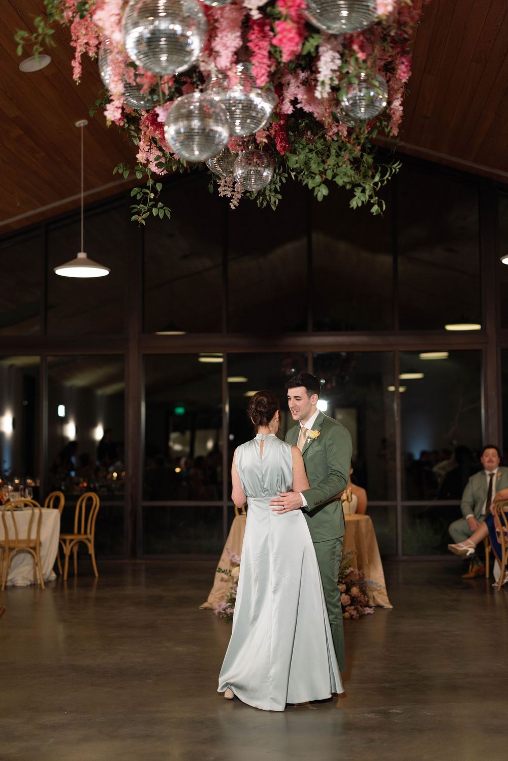 A couple shares their first dance under disco balls and pink floral decorations in an elegant reception venue.
