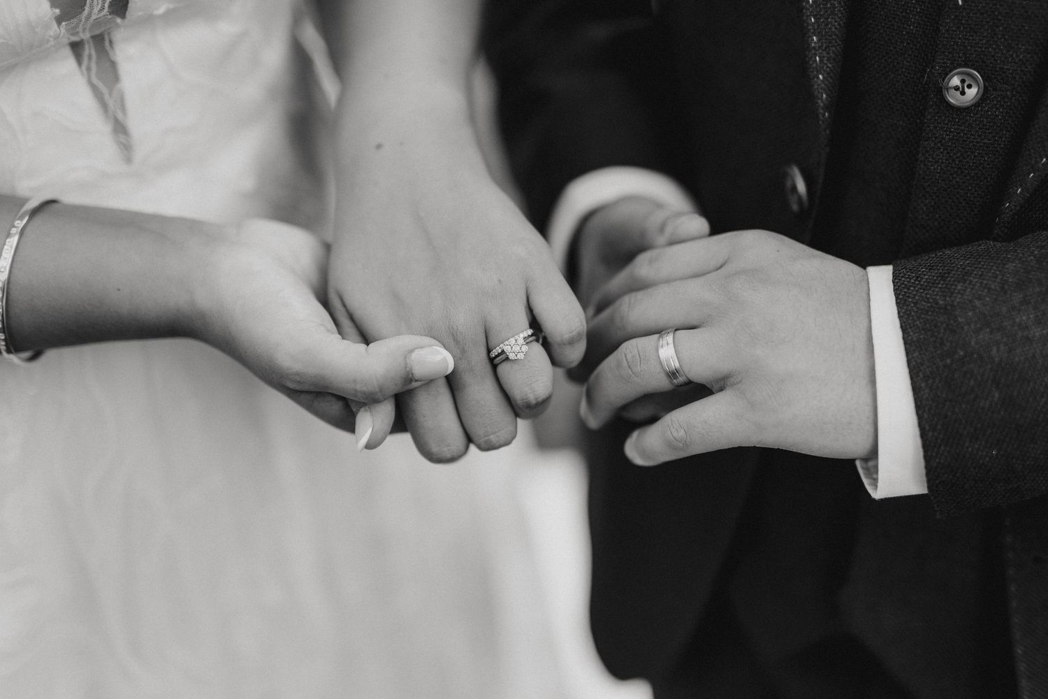 A black and white close-up photograph shows hands exchanging wedding rings during a marriage ceremony.