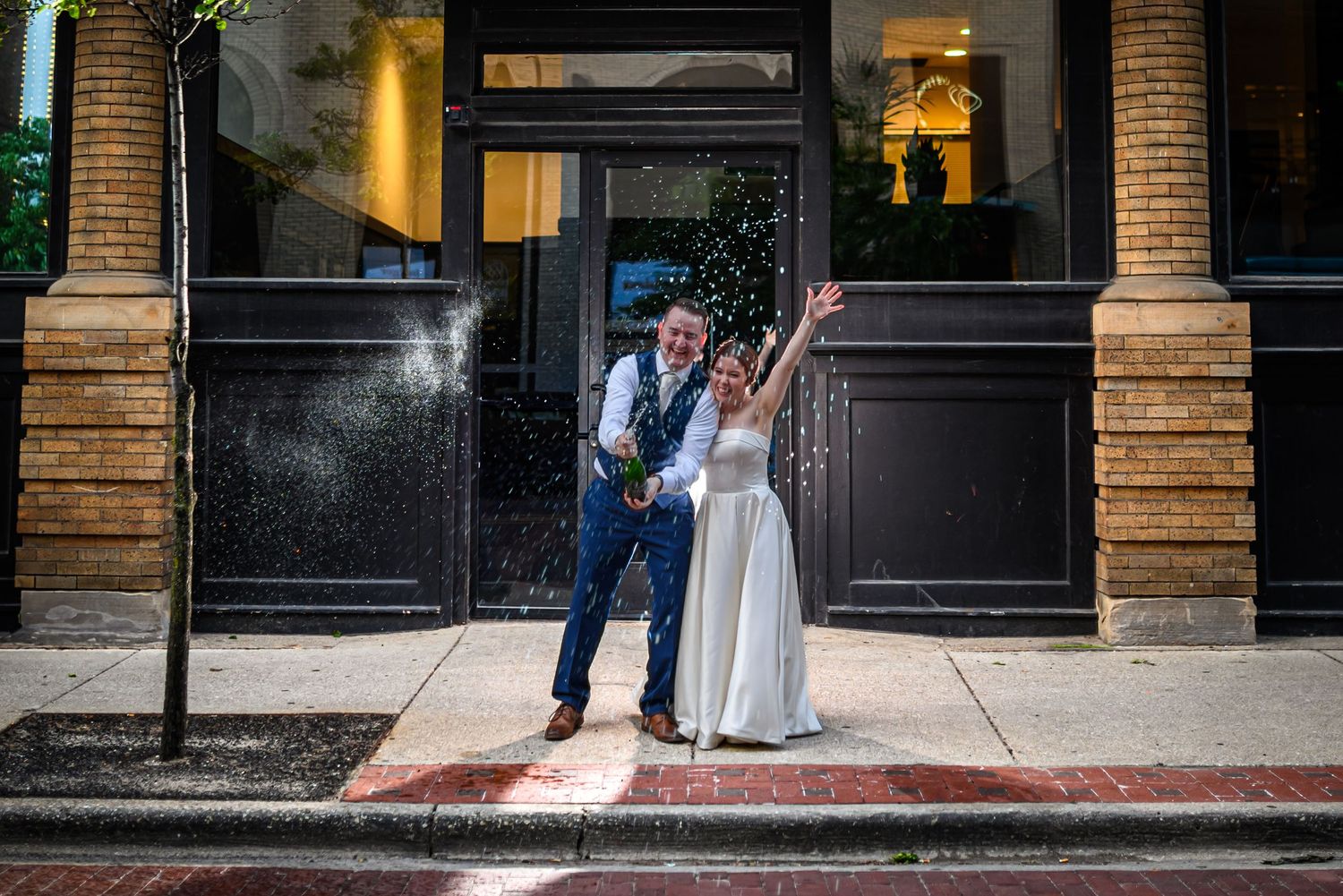 A couple in wedding attire celebrates joyfully outside a building with brick columns and large windows.