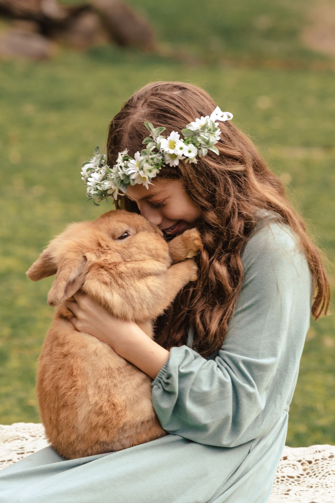 A person in a light blue dress embraces a fluffy brown bunny rabbit while sitting outdoors on a sunny day.