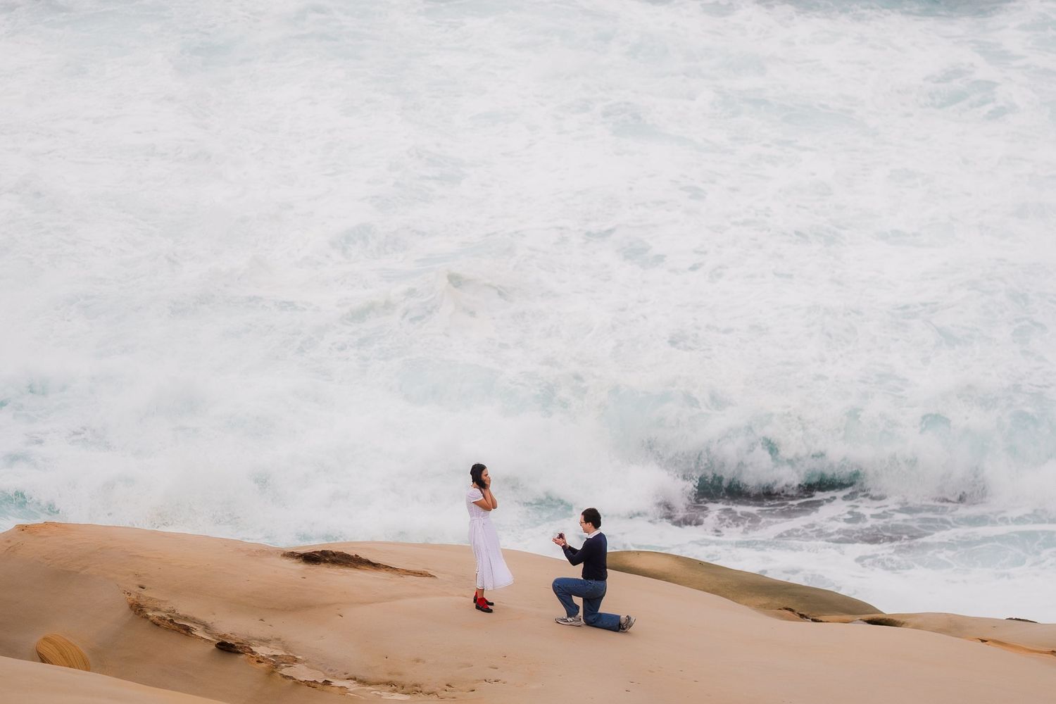 Aerial view of crashing ocean waves against rocky coastline with people visible on sandy cliffs at sunset.