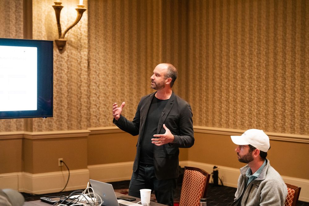 Speaker presenting to an audience in a conference room with warm lighting and striped wallpaper.