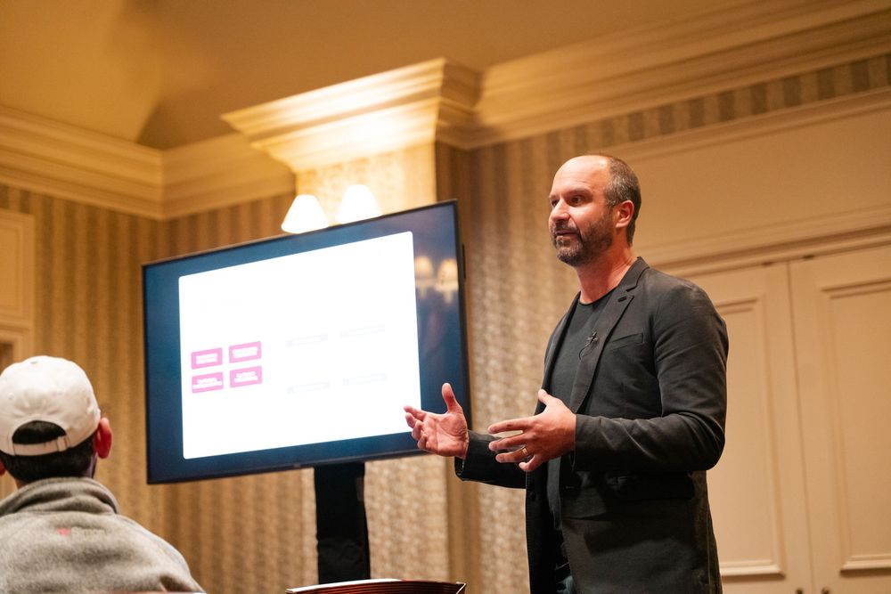A speaker gestures while giving a presentation in a conference room with white walls and decorative molding.