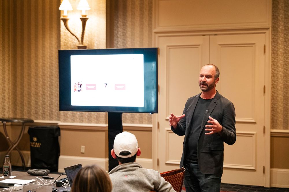 Presenter gestures while speaking at a professional conference in an elegant meeting room with wall sconces.