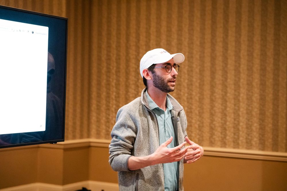 A person in a white baseball cap gestures while speaking at a podium during a presentation.