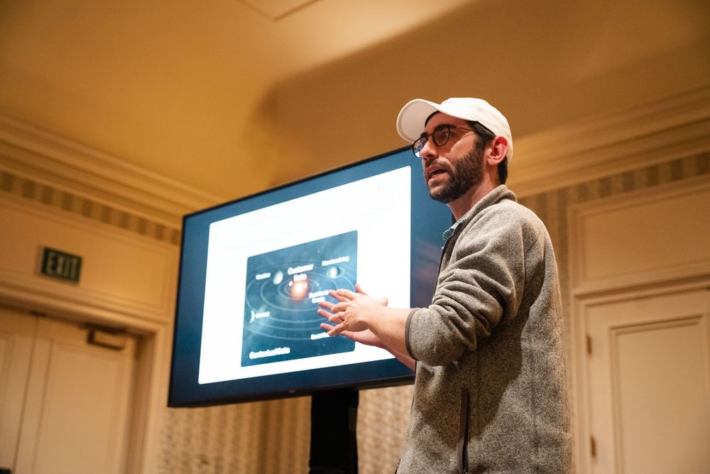A speaker presents to an audience in an elegant room with multiple display screens mounted on the walls.