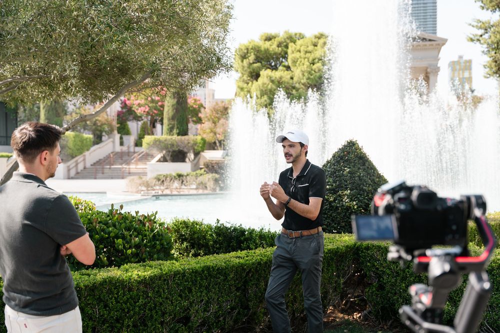 Two people share a romantic moment in front of a garden fountain during a sunny afternoon.