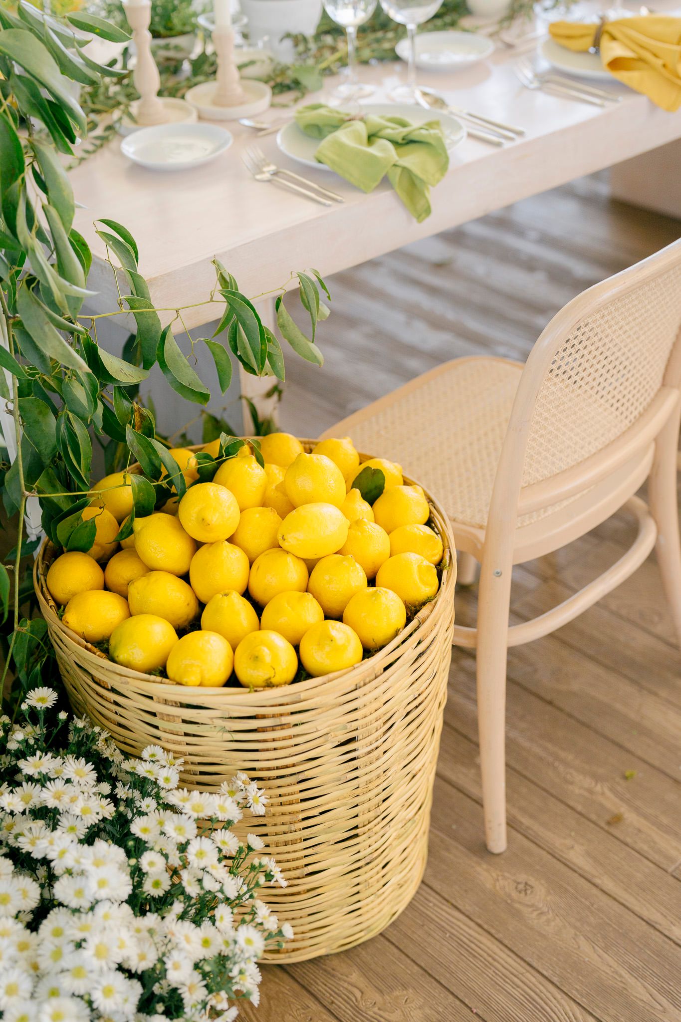 Fresh yellow lemons fill a woven basket on a wooden deck with delicate white flowers and greenery as decoration.