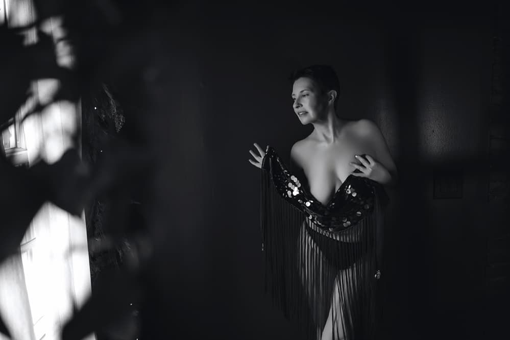 Dramatic black and white boudoir photograph with soft window lighting creating shadows and silhouettes.