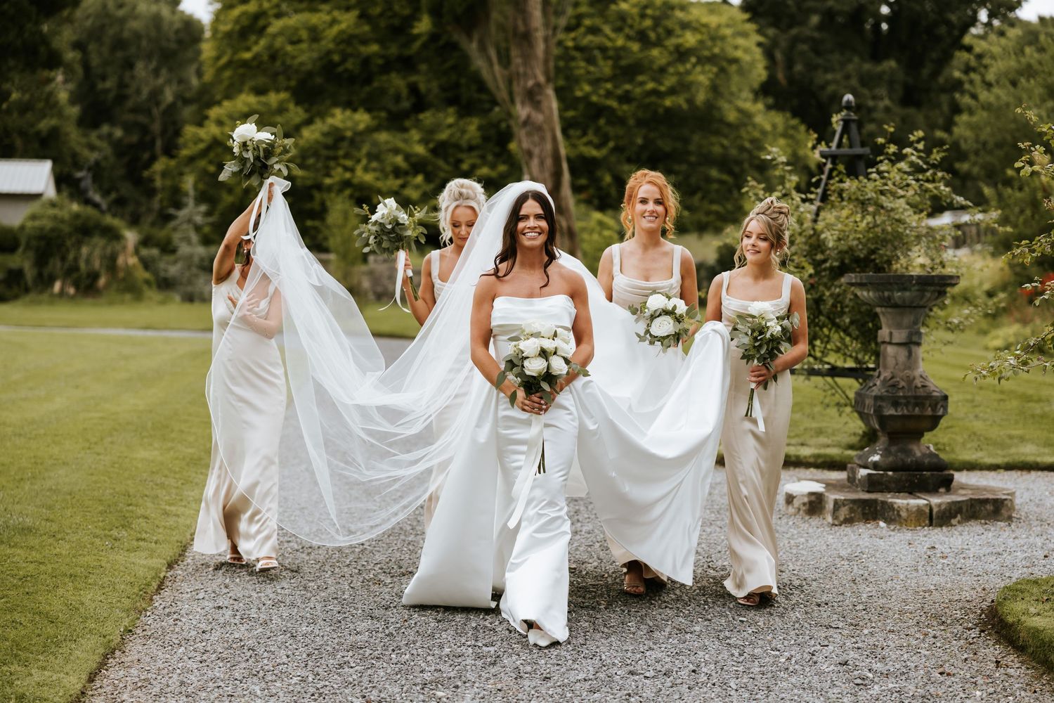 Bridesmaids in flowing white dresses walk together holding bouquets in garden during a sunny wedding. Markree Castle Conlon Photo&Film